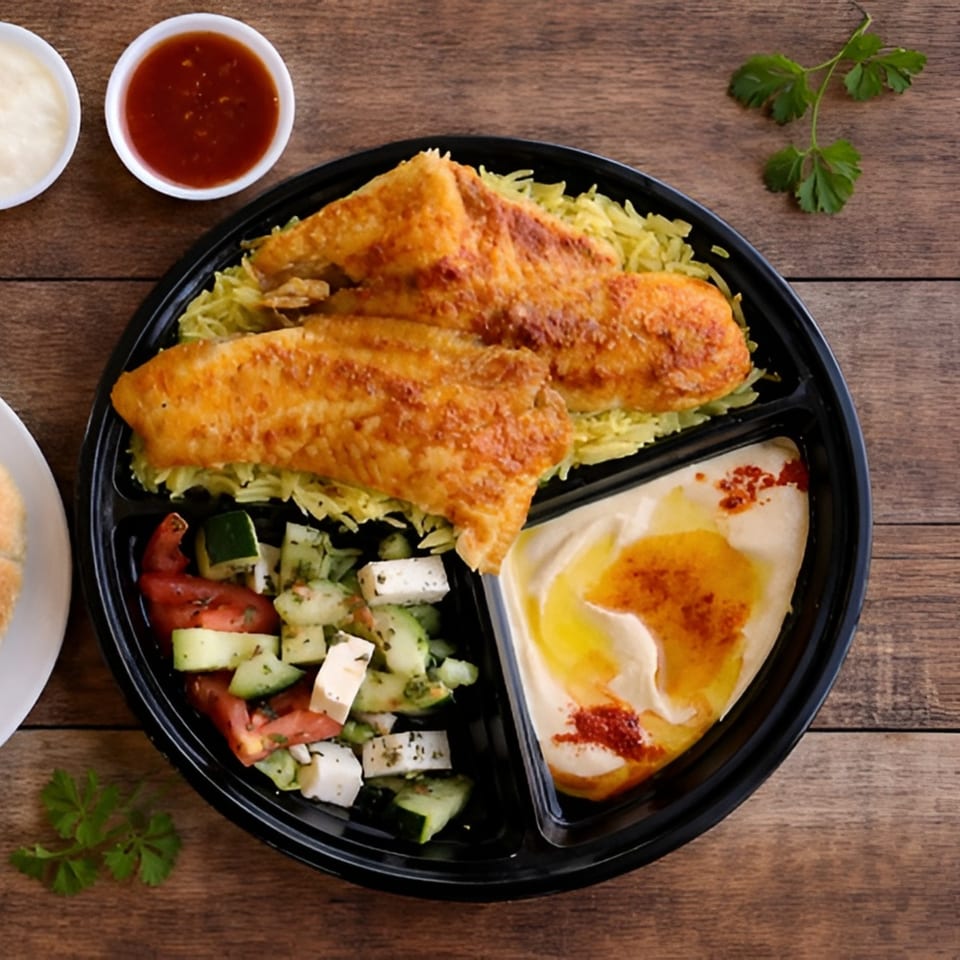 A takeout container with fried fish over rice, a side salad, and hummus on a wooden table.