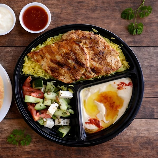 Grilled chicken, yellow rice, hummus, and a cucumber and tomato salad in a black to-go container.