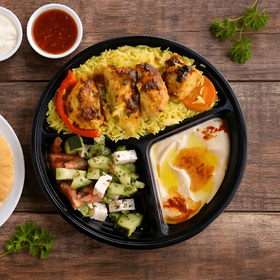 A black partitioned plate with chicken and rice, a cucumber and feta salad, and hummus.