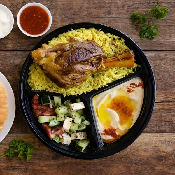 Lamb shank with yellow rice, salad, hummus, and sauces in a black divided plate.