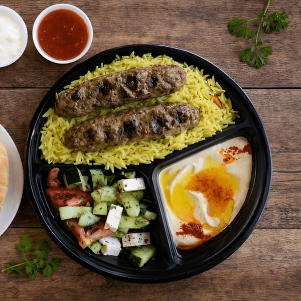 A black divided plate with two grilled kebabs on yellow rice, a cucumber and tomato salad, and hummus.